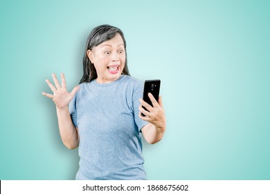 Portrait of shocked old woman reading a news on her mobile phone while standing in the studio. Isolated on white background
