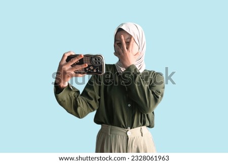 portrait of shocked, scared asian muslim woman peeking between her fingers at cell phone isolated on white background