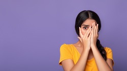 Portrait Of Shocked And Scared Young Indian Lady Covering Her Eyes With Palms And Peeking Through Fingers, Standing Isolated Over Purple Studio Background, Banner, Free Copy Space. Phobia Concept