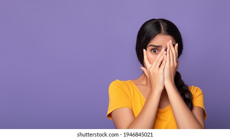 Portrait of shocked and scared young indian lady covering her eyes with palms and peeking through fingers, standing isolated over purple studio background, banner, free copy space. Phobia Concept