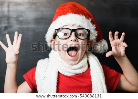 Portrait of a shocked small boy in Santa Claus hat black glasses red shirt and white knitted scarf