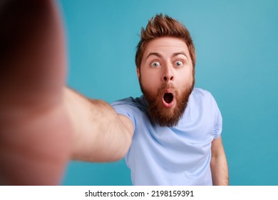 Portrait of shocked surprised handsome young adult bearded man standing with widely open mouth, making selfie POV, expressing astonishment. Indoor studio shot isolated on blue background. - Powered by Shutterstock