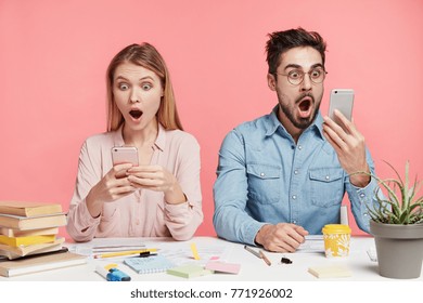 Portrait of shocked terrified male and female stare at mobile phones, type messages or read shocking news, sit next to each other at work place, isolated over pink background. Suprised students indoor