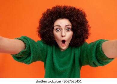 Portrait of shocked woman with Afro hairstyle wearing green casual style sweater standing with open mouth, making selfie POV, expressing astonishment. Indoor studio shot isolated on orange background.