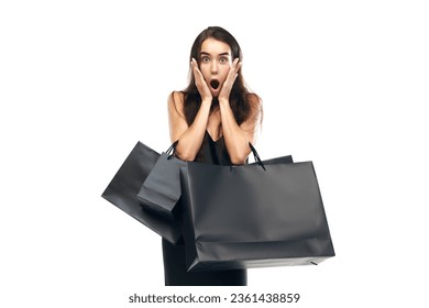 Portrait of shocked woman wearing black dress raising hands with bags. Beginning season of sales. Concept of Black Friday, Cyber monday, fashion, buying, holiday shopping. Copy space for ad