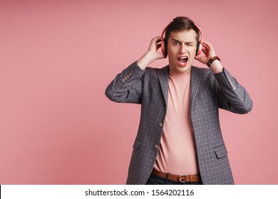 Portrait of a shocked young attractive man wearing jacket standing isolated over pink background, lstening to loud music with wireless headphones