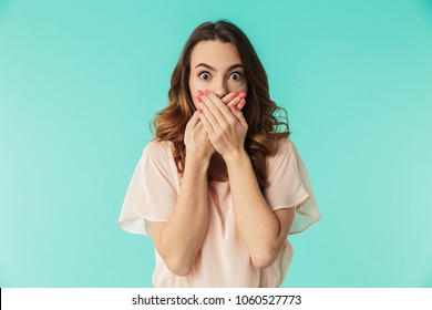 Portrait of a shocked young girl in dress looking at camera with mouth covered isolated over blue background