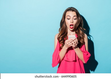 Portrait if a shocked young girl in dress looking at mobile phone isolated over blue background - Powered by Shutterstock