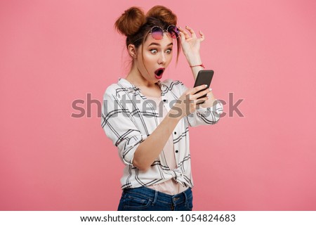 Portrait of a shocked young girl using mobile phone isolated over pink background