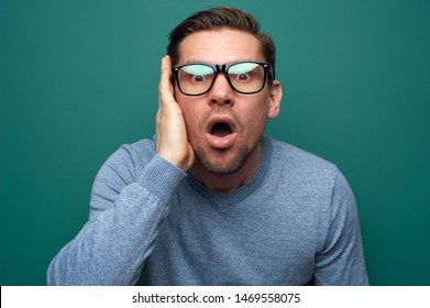Portrait of a shocked young man in glasses and a blue shirt posing on a green background with a hand near his face. The concept of shock and surprise. Advertising space