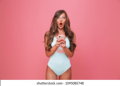 Portrait of a shocked young woman dressed in swimsuit holding mobile phone isolated over pink background