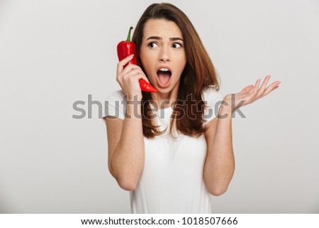 Portrait of a shocked young woman holding chili pepper at her ear and calling isolated over white background