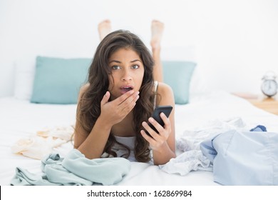 Portrait Of A Shocked Young Woman With Mobile Phone In Bed At Home