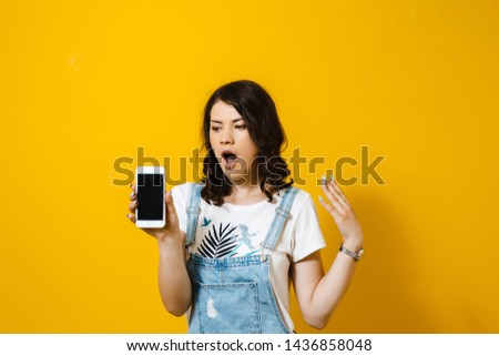 Portrait of a shocked young woman showing blank screen mobile phone isolated over yellow background