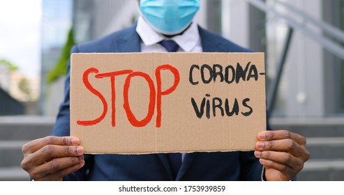 Portrait shot of African American young man in medical mask standing outdoor at big stairs and showing table with words Stop Coronavirus. Male demonstrating board with protest against pandemic. - Shutterstock ID 1753939859