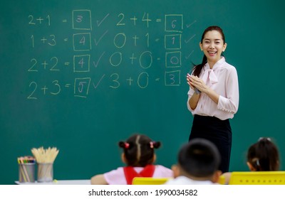 Portrait Shot Of Asian Beautiful Female Mathematic Teacher Standing Smiling Cheer Up While Little Smart Schoolgirl Student Solving Math Equations On Chalkboard In Front Of Classmates In Classroom.