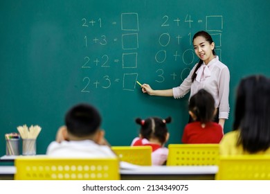 Portrait shot of Asian beautiful female mathematic tutor standing smiling holding chalk writing elementary school math equation questions on chalkboard while group of boy and girls students studying.