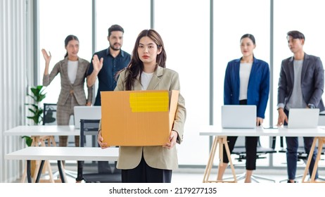 Portrait Shot Of Asian Sad Jobless Businesswoman In Casual Suit Standing Holding Belongings In Cardboard Box After Fired While Male And Female Colleagues Waving Hands Goodbye In Blurred Background.