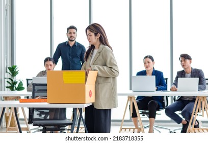 Portrait Shot Of Asian Sad Jobless Businesswoman In Casual Suit Standing Holding Belongings In Cardboard Box After Fired While Male And Female Colleagues Waving Hands Goodbye In Blurred Background.