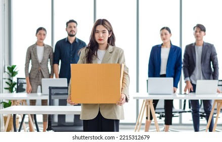 Portrait Shot Of Asian Sad Jobless Businesswoman In Casual Suit Standing Holding Belongings In Cardboard Box After Fired While Male And Female Colleagues Waving Hands Goodbye In Blurred Background.