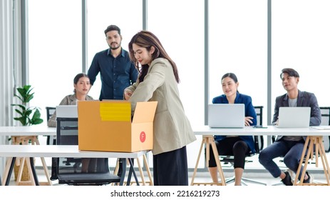 Portrait Shot Of Asian Sad Jobless Businesswoman In Casual Suit Standing Holding Belongings In Cardboard Box After Fired While Male And Female Colleagues Waving Hands Goodbye In Blurred Background.