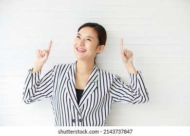 Portrait shot of Asian young confident happy female businesswoman wears black white stripe casual suit stand in front wall smiling hold hands point fingers and look up above on empty copy space.