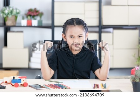 Portrait shot of Asian young fierce serious elementary school girl in casual outfit sitting look at camera holding fists up showing strength biceps arms at working desk with smartphone tablet and toy.