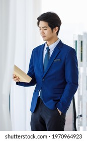 Portrait Shot Of Asian Young Intelligence College Teen Law Student Wears Blue Formal Suit Uniform And Necktie Standing Hold And Read Textbook Cover In Hand In Front Bookshelf In University Library.