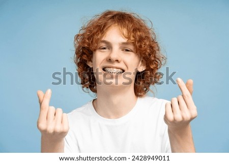 Portrait shot of an attractive curly haired redhead male teen, smiling with braces and making a Korean love gesture at the camera, set against a blue studio background