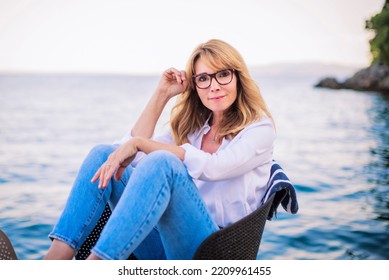 Portrait Shot Of Attractive Mature Woman Wearing White Shirt And Blue Jeans While Sitting On The Chair And Relaxing By The Sea.