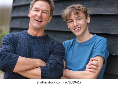 Portrait Shot Of An Attractive, Successful And Happy Middle Aged Man Father Male Arms Folded With His Teenage Boy Son Outside Laughing Teen, Wearing A Blue Sweater And T-shirt