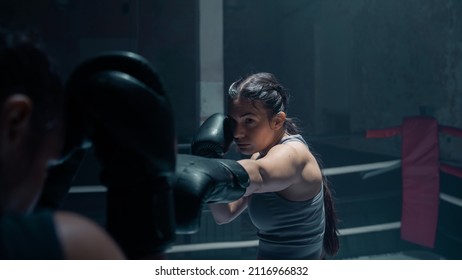 Portrait Shot Of A Badass Confident Looking Ethnic Female Boxer Training With Her Coach, Hitting The Pads As She Protects Her Face. Background Is A Beat Down Old Boxing Gym Studio. Martial Arts Women.