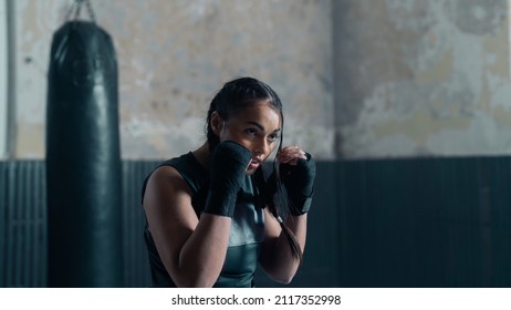 Portrait Shot Of A Badass Female Kickboxer With Braids Training In An Old Boxing Gym Studio, Wearing Gloves And A Tank Top, Punching Bag Behind Her. Confident Boxer Practising Sparring Alone.