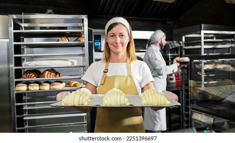Portrait shot of beautiful female in white apron and hat standing in front of camera, holding tray with fresh just-baked croissants in kitchen of bakehouse. Woman in bakeshop showing pastry. - Shutterstock ID 2251396697