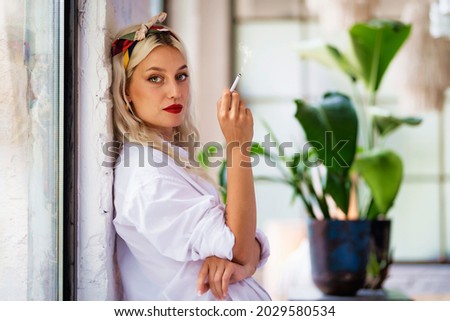 Portrait shot of beautiful green eyed young woman wearing hair scarf and white shirt while smoking cigarette by the window at home.