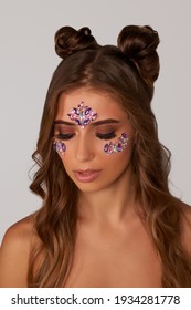     Portrait shot of brown-haired lady with two hair buns and shining pink and violet crystal patterns on forehead and under eyes. Temporary tattoos are made as waterproof non-toxic skin stickers.    