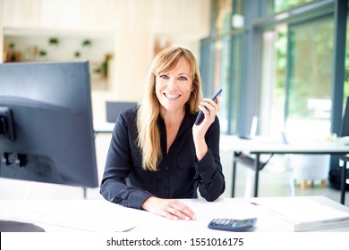 Portrait shot of businesswoman withh toothy smile sitting at office desk and holding mobile phone in her hand.