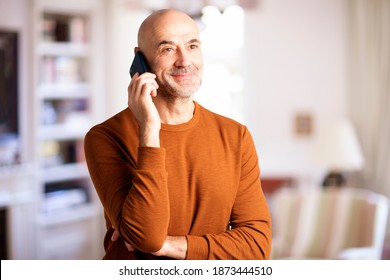 Portrait Shot Of Casual Man Talking With Somebody On His Mobile Phone While Standing In The Living Room At Home. 