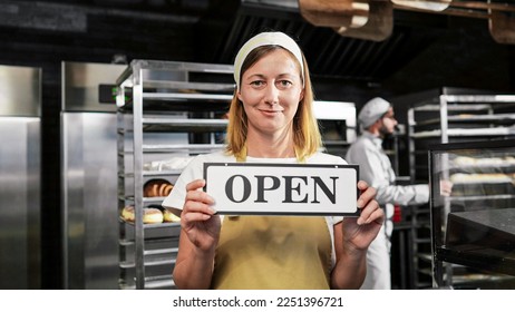 Portrait shot of Caucasian beautiful female baker in apron and hat standing in bakehouse in the morning and holding table Open. Bakery opening early concept. Good-looking woman chief in cafe. - Shutterstock ID 2251396721