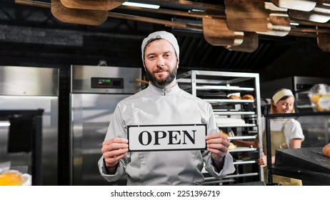Portrait shot of Caucasian handsome male baker in apron and hat standing in bakehouse in the morning and holding table Open. Bakery opening early concept. Good-looking man chief in cafe. - Shutterstock ID 2251396719