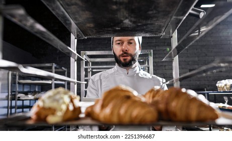 Portrait shot of Caucasian handsome man in hat and uniform working in bakehouse and checking croissants. Male baker with beard making fresh pastries in kitchen of bakeshop in morning. - Shutterstock ID 2251396687