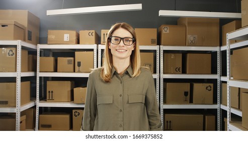 Portrait Shot Of Caucasian Happy Beautiful Young Woman In Glasses Standing In Postal Store With Carton Boxes On Shelves. Female Post Worker Smiling Joyfully To Camera In Mail Office Among Parcels.