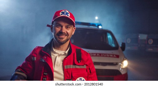 Portrait Shot Of Caucasian Happy Smiled Handsome Man Medic Taking On Hat And Smiling To Camera. Ambulance Car On Background. Cheerful Attractive Male Paramedic At Night Shift.