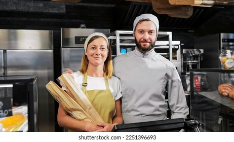 Portrait shot of Caucasian joyful couple of male and female bakers standing at bakehouse kitchen with baguettes. looking at each other and smiling to camera. Happy man and woman workers of bakeshop. - Shutterstock ID 2251396693