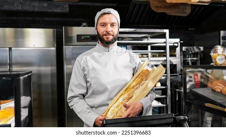 Portrait shot of Caucasian young handsome male baker in uniform standing at bakehouse kitchen with baguettes and smiling. Happy man worker looking at camera on bakeshop. Fresh bread in the morning. - Shutterstock ID 2251396683