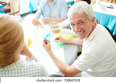 Portrait Shot Of Cheerful Senior Man Looking At Camera With Toothy Smile While Making Origami Figure From Colorful Paper, His Friends Sitting Next To Him