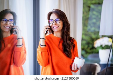 Portrait shot of cheerful woman making a phone call while standing at the window at home.  - Powered by Shutterstock