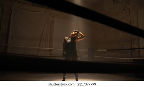 Portrait Shot of a Confident Badass Female Kickboxer Standing in the Middle of a Boxing Ring With Light Shining on Her From the Top As She is Stretching and Warming Up Her Body To Start Training. - Powered by Shutterstock