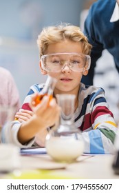 Portrait Shot Of A Cute Little Boy With Safety Glasses Mixes Chemicals In Beakers. Elementary School Science Classroom. Children Learn With Interest