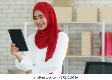 Portrait shot of cute smiling young confident Muslim woman wearing a red hijab standing at her office, looking at camera while holding a digital tablet with boxes and paper bags background - Powered by Shutterstock